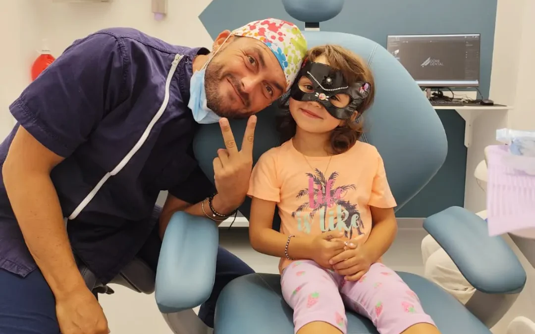 Bambina sorridente sulla poltrona del dentista con una maschera da gatto, accanto al dentista pediatrico, durante un controllo per i denti da latte