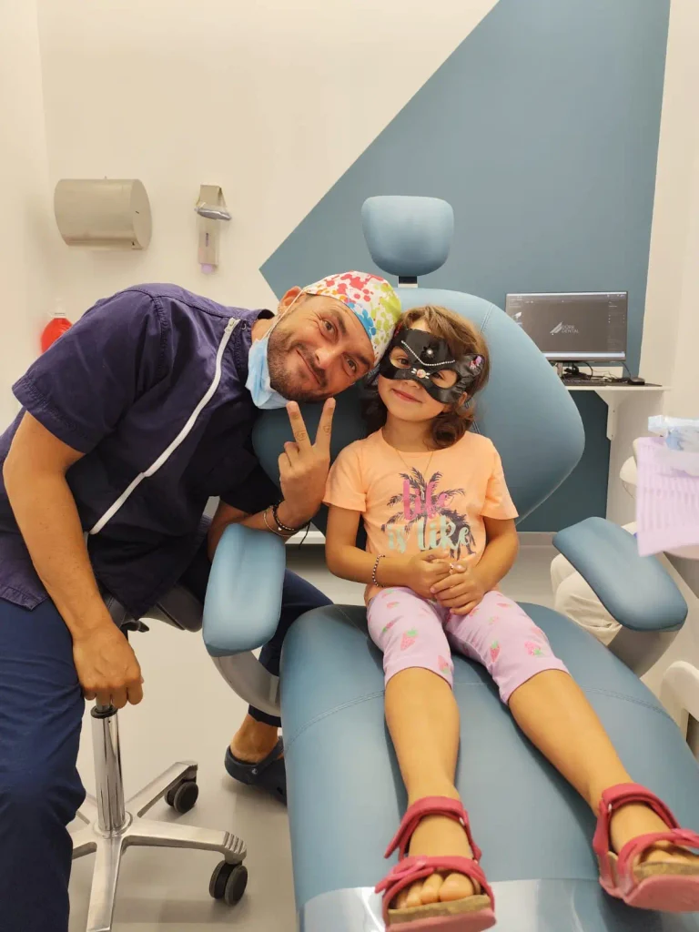 Bambina sorridente sulla poltrona del dentista con una maschera da gatto, accanto al dentista pediatrico, durante un controllo per i denti da latte.